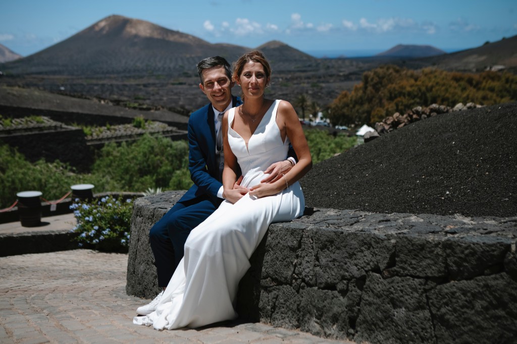 Bride and groom sitting on a stone fence in La Geria, captured by a wedding photographer in Lanzarote, perfect for elopements