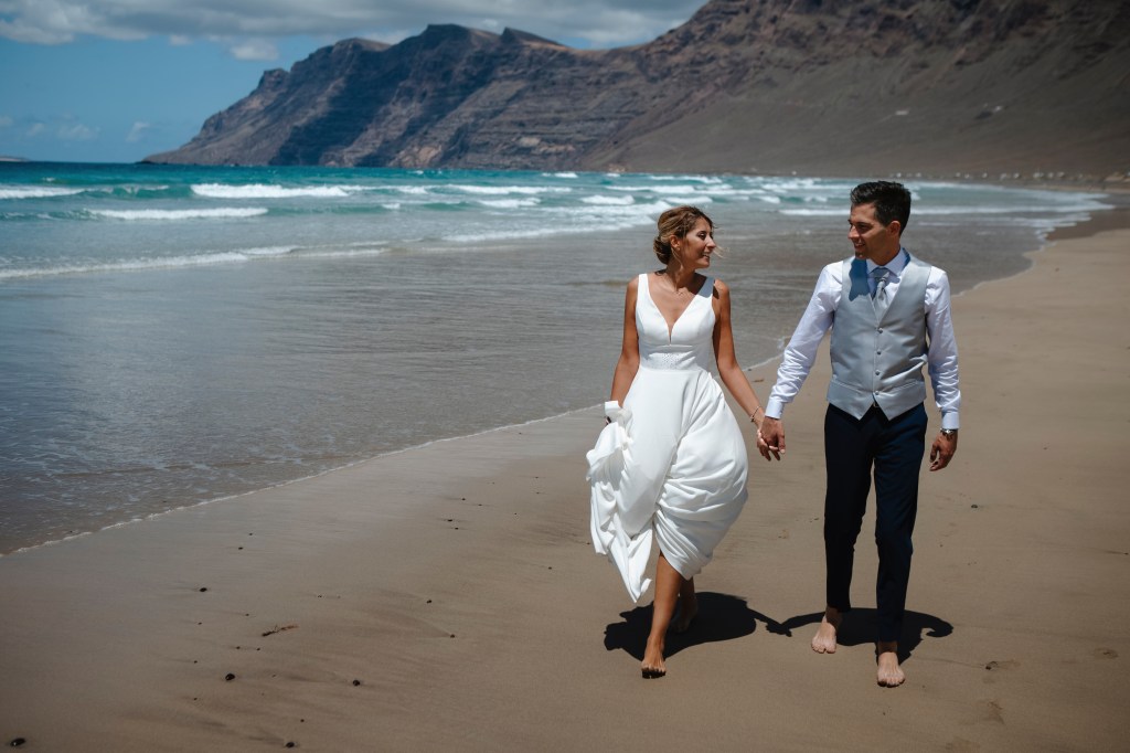 Bride and groom walking towards wedding photographer on Famara Beach, Lanzarote, capturing their elopement against a stunning landscape