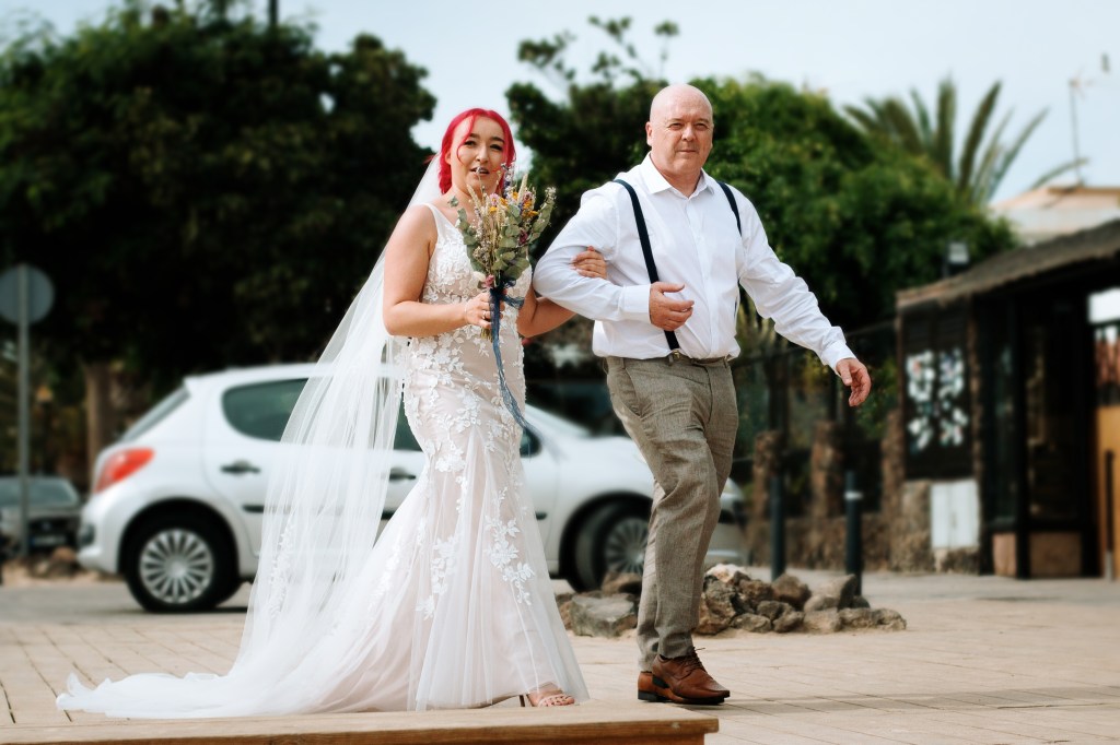 Father walks bride to her wedding ceremony in Corralejo and both are looking to Fuerteventura wedding photographer