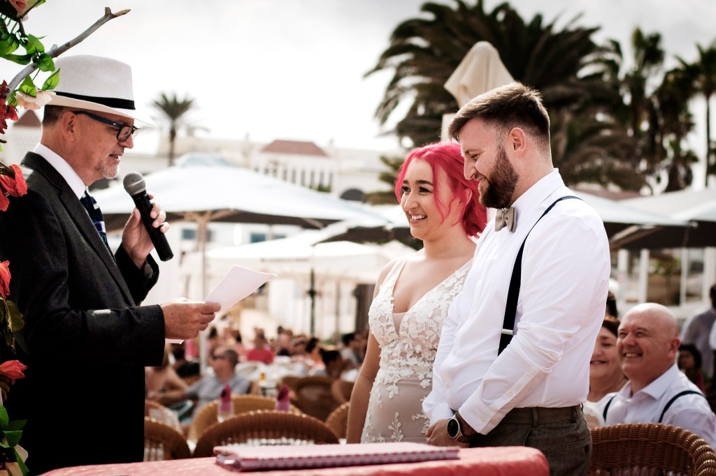 Emotional vow exchange in Corralejo - bride and groom hold hands under floral arch as celebrant officiates with beach guests watching