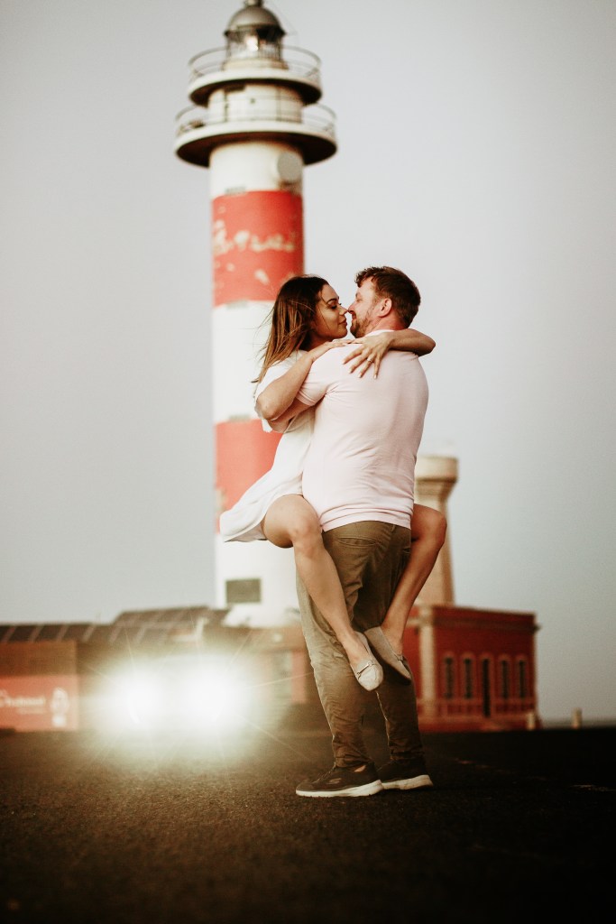 Professional couple photography in Fuerteventura and Lanzarote: Loving couple shares a kiss as girl jumps on her boyfriend near El Cotillo lighthouse