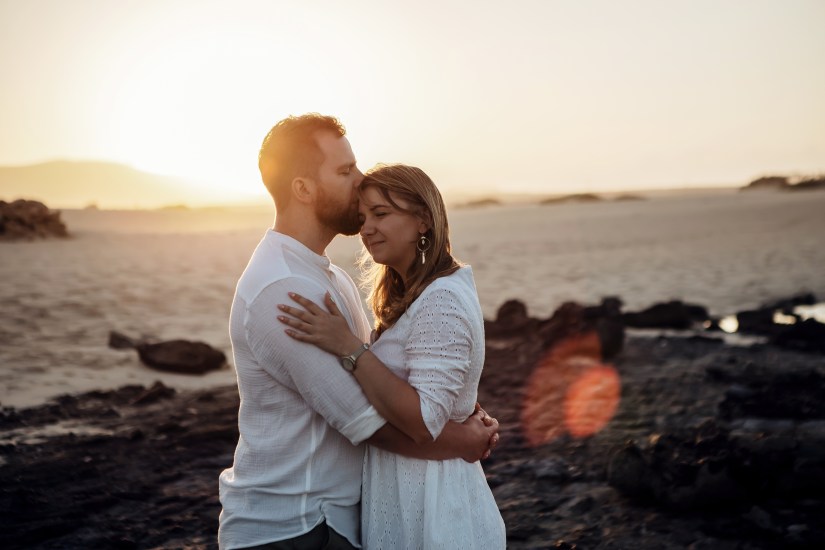 Fuerteventura and Lanzarote Couple Photographer captures a couple during golden hour in the Dunes of Corralejo, with the sun shining behind them. The couple exchanges loving glances, as the photographer captures this romantic moment in the beautiful landscapes of Fuerteventura and Lanzarote.