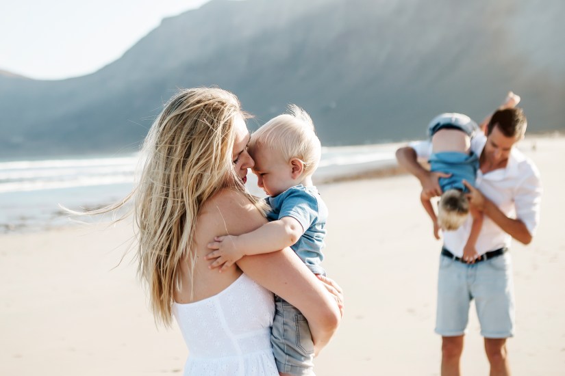 Frohe Familie, die den wunderschönen Strand von Famara auf Lanzarote genießt, spritzt im Wasser, während die Sonne durch den Berg im Hintergrund scheint. Aufgenommen von einem erfahrenen Familienfotografen auf Lanzarote.